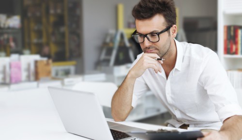 Парень сидит за столом напротив ноутбука и держит в руках ручку